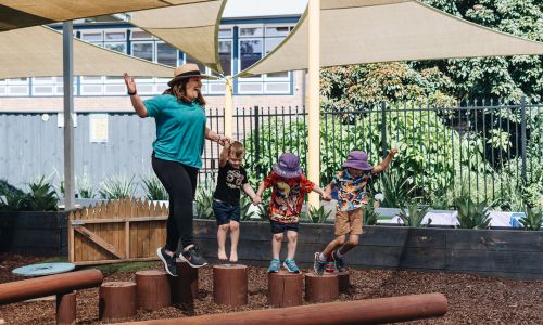 An educator holding hands with three children as they balance along timber logs in the outdoor play yard at Hornsby.