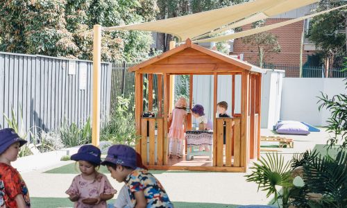 Children in sun hats playing beside a wooden cubby house on the synthetic grass playground.