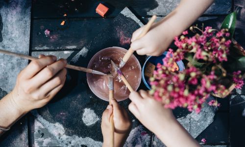 Children's hands painting with brushes around a glass bowl and native pink blossoms on a dark table.