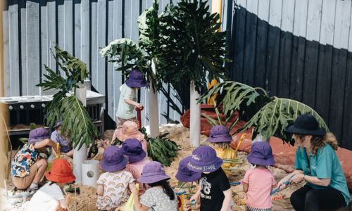 A group of children and an educator digging together in the shaded sandpit surrounded by tropical planting.