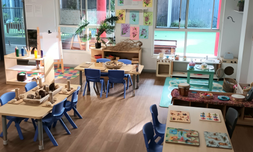 The toddler room with timber shelving, a home corner and small tables set with play materials.