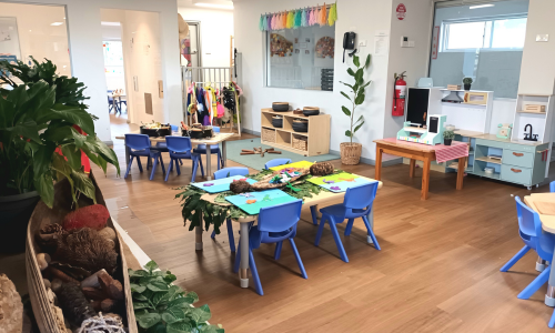A bright classroom with blue chairs at low tables, a play kitchen and plenty of natural light.